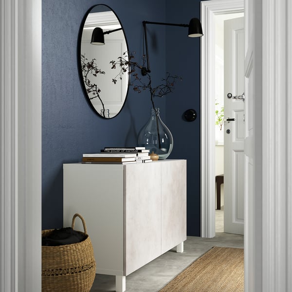 Diagonal vy av modern hall med blå vägg, vit BESTÅ-skänk, stor spegel, korg, böcker, vas och vägglampor.