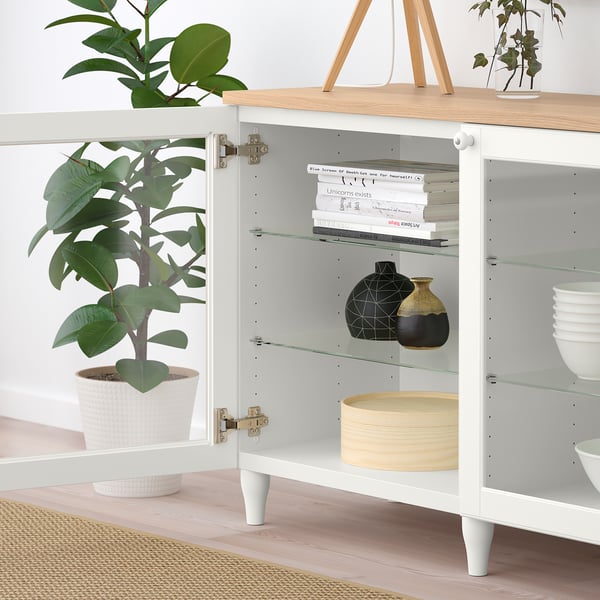 Bilden visar ett vitt BESTÅ sideboard med glasdörrar, som visar hyllor med böcker, vaser, skålar och krukväxter på ett trägolv.