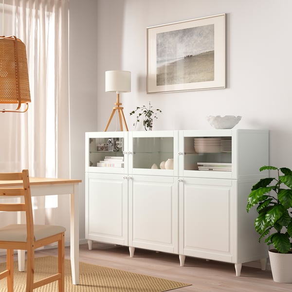 Modernt BESTÅ sideboard med glasdörrar och lådor, bredvid en trästol, med inredning och böcker.