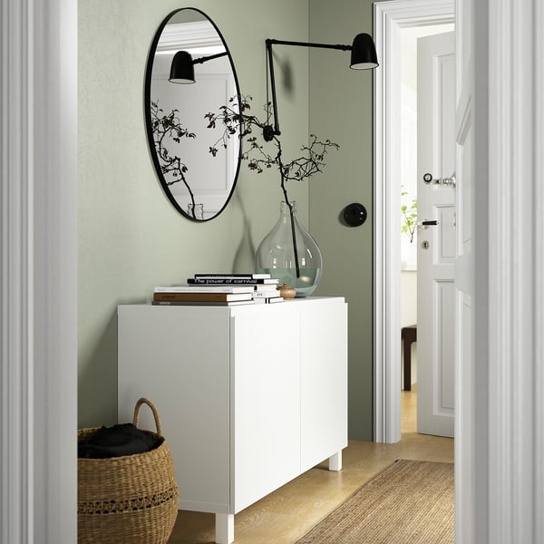 Modern hall med BESTÅ vit skänk, bred och låg, rymmer böcker, vas med grenar och spegel ovanför. Korg nedanför, dörrar öppnas till ett annat rum.