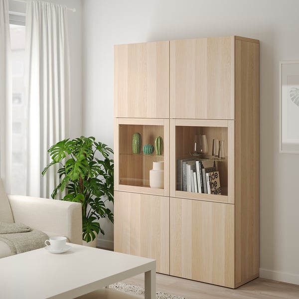 Ikea BESTÅ förvaringsskåp i ljus ek med glasdörrar och blandade hyllor bredvid soffbord och soffa.