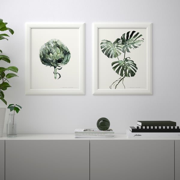 Två inramade botaniska tryck hänger ovanför en grå skänk, som rymmer en växt, vas, bok, tidskrift och dekorativ sfär.