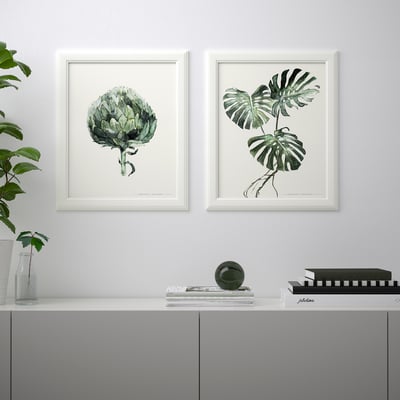 Två inramade botaniska tryck hänger ovanför en grå skänk, som rymmer en växt, vas, bok, tidskrift och dekorativ sfär.