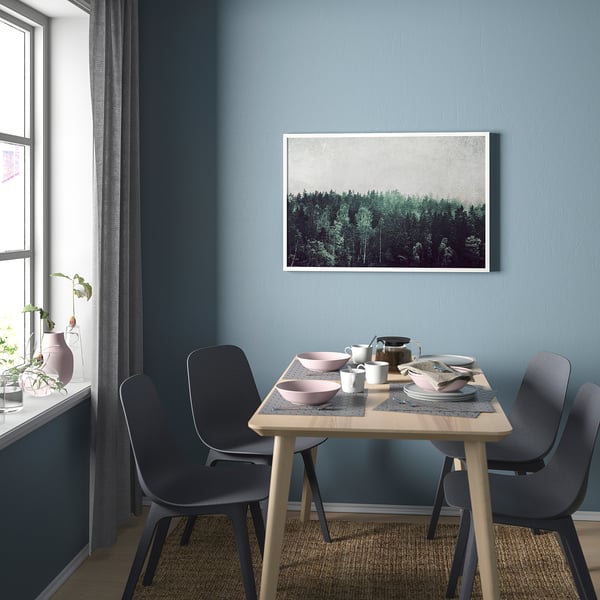 En modern matplats med ett träbord och svarta stolar. Den blå väggen har en inramad skogsaffisch. Ett fönster med en blomvas finns.