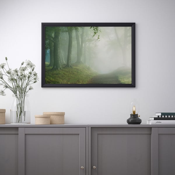 En inramad naturscen ovanför ett grått skåp, med blommor, askar och en lampa.
