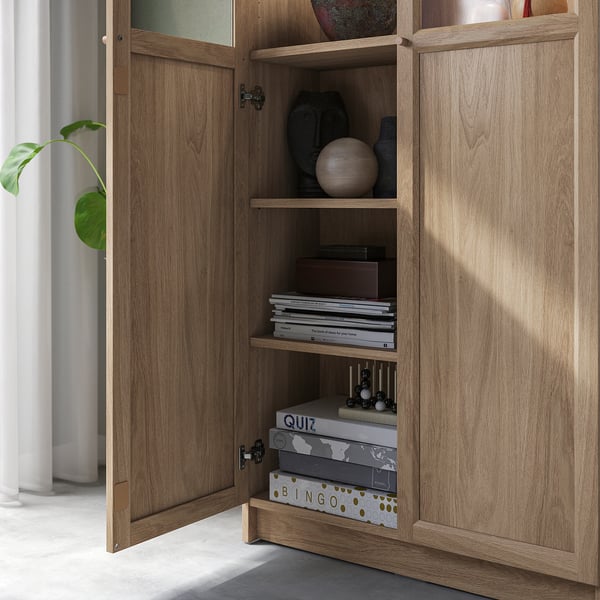 Bokhylla i trä med glasdörrar som visar hyllor fyllda med böcker, inredningsartiklar och spel, vilket visar mångsidiga förvarings- och visningsmöjligheter.