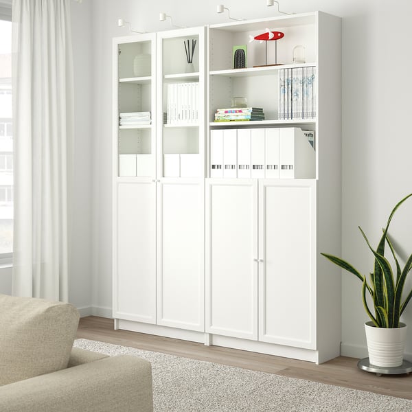 Vit BILLY-bokhylla med glasdörrar, som rymmer böcker och inredning, bredvid en beige stol och en stor krukväxt.