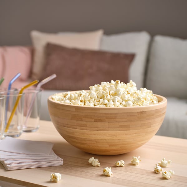 En stor träskål fylld med popcorn står på ett soffbord nära glas och kuddar.
