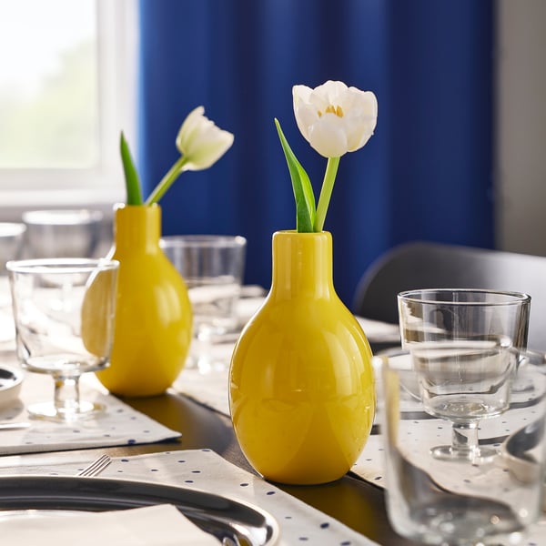 Tre gula vaser rymmer enstaka vita blommor på ett bord dukat med glas och blåprickiga servetter.