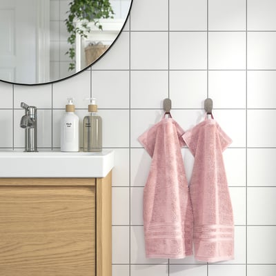 BROKGLIM rosa handdukar hänger snyggt på kromkrokar bredvid ett modernt handfat med vit bänkskiva, spegel och vedskåp. Mjuk, absorberande och ekologisk.