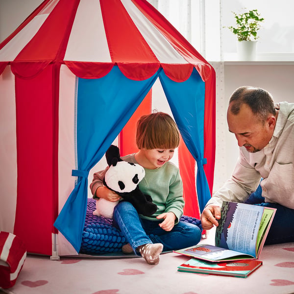 Ett barn sitter i ett färgglatt lektält med en man som läser en bok och illustrerar ett mysigt gömställe där barnen kan leka eller lyssna på sagor.