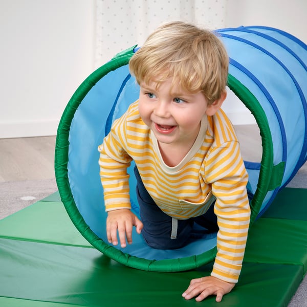 Ett barn leker inuti en färgglad plasttunnel och kryper genom den på en grön matta.