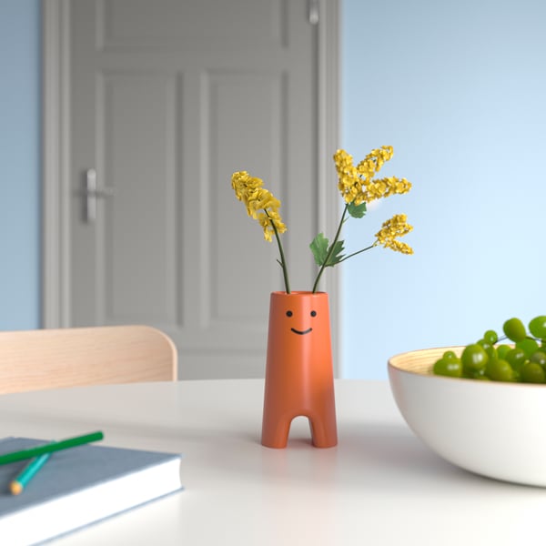 Orange FRÖVÄXT vas med leende ansikte, liten, rymmer gula blommor, på vitt bord vid skål med druvor.