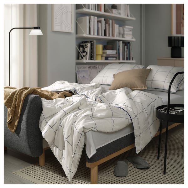 Modernt sovrum med obäddad bäddsoffa GRUNNARP, vita gallersängkläder, solbrända kuddar, grå tofflor, bokhylla och golvlampa.