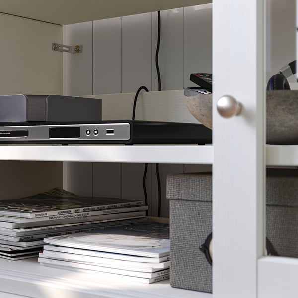 Vit trähylla med elektronik och tidningar. Fjärrkontroll på toppen, kablar synliga.