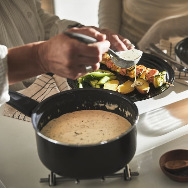 Person som serverar spett grönsaker från svart tallrik bredvid krämig kastrull på bänken.