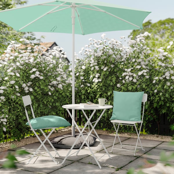 Litet utomhusbord med ljusgröna vadderade stolar och myntaparaply, omgivet av blommande buskar.