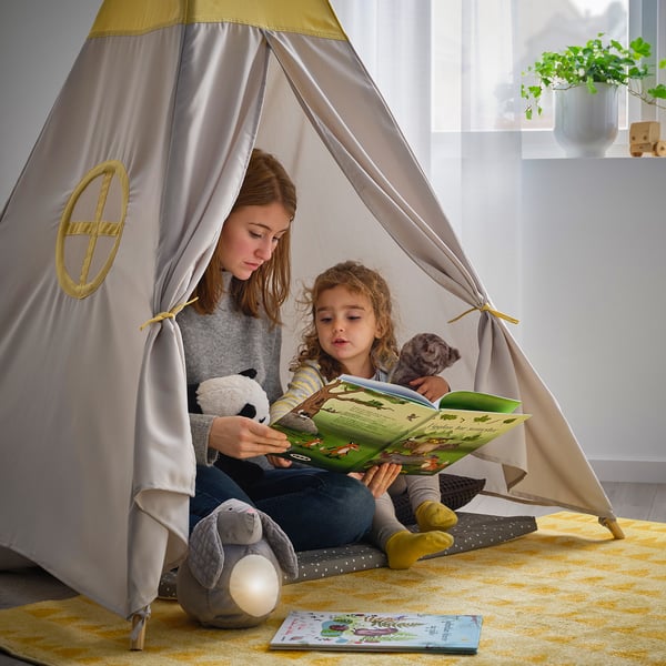 Ung flicka läser för småbarn inuti beige och gult tält. Gosedjur gör dem sällskap.