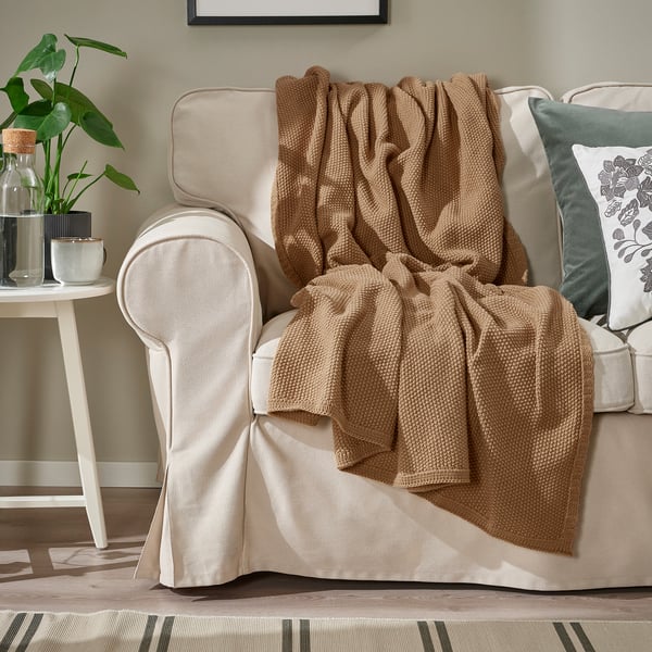 En beige filt är draperad över en krämfärgad fåtölj bredvid ett sidobord med en växt.