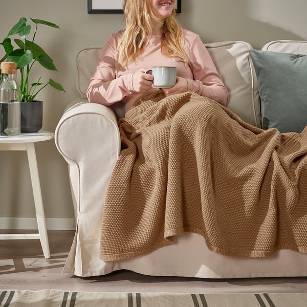 Kvinna som håller muggen, insvept i mjuk brun HUMLEMOTT pläd på beige stol.