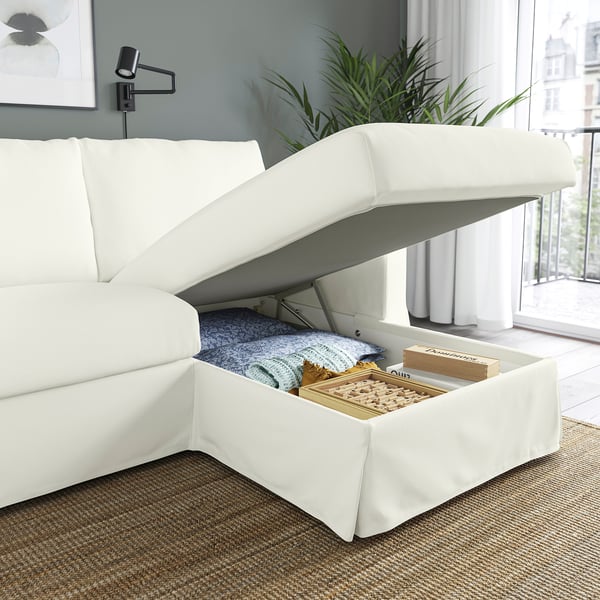 En modern vit soffa med en uppfällbar schäslong som avslöjar brädspel och filtar i förvaringen.