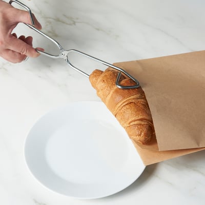 Tång som plockar upp ett bakverk från en brun papperspåse över en vit skål.