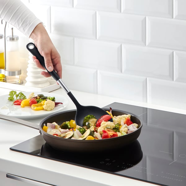 Person som lagar färgglada grönsaker i en kastrull med en bred, non-stick spatel med ett greppvänligt handtag på en modern spis.