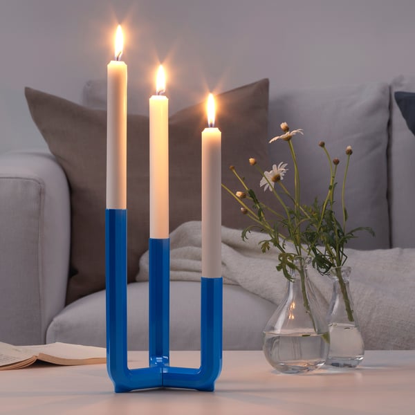 Blå ljusstake med tre tända vita ljus sitter på ett bord, bredvid två små glasvaser med blommor, med en beige soffa i bakgrunden.