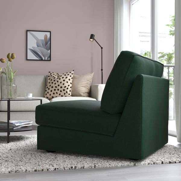 Modern grön bäddsoffa, rosa väggar, grå soffa med kuddar, lampa, bokhylla och fönsterutsikt.