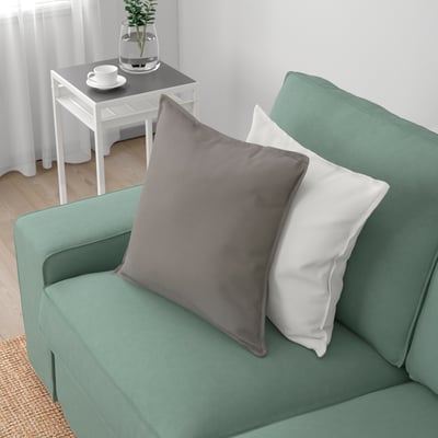 En mysig grön soffa med grå och vita kuddar sitter i ett ljust rum och visar upp sina bekväma, djupa kuddar och robusta design, vilket inbjuder till avkoppling.