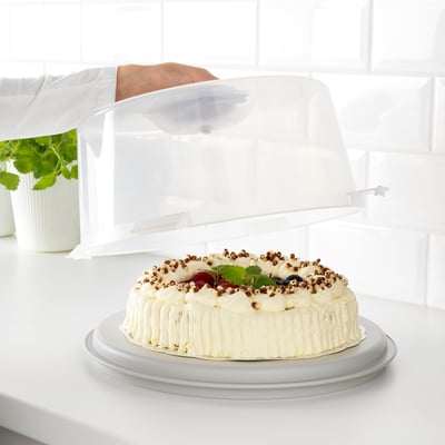 En tårta i en bärare med genomskinligt lock, placerad på en bänkskiva. Bäraren har räfflor och släta sidor för olika bakverk.