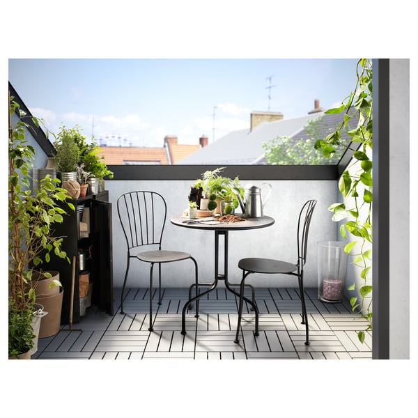 Liten balkong med svart metallbord och två stolar, omgiven av krukväxter. Mysig urban utomhusmiljö.