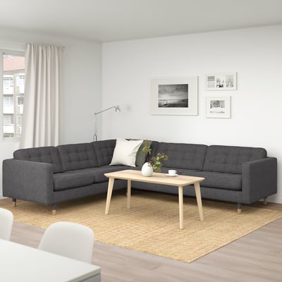 Modernt vardagsrum med stor, mörkgrå l-formad sektionssoffa som mittpunkt, ljust soffbord i trä, beige matta och väggfoton.