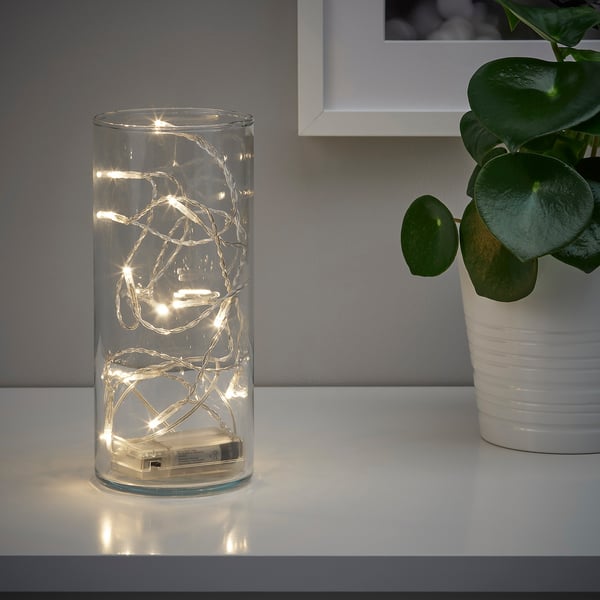 En klar vas håller varmvita LED-lampor och skapar en mysig atmosfär. En krukväxt står bredvid den på en vit yta.