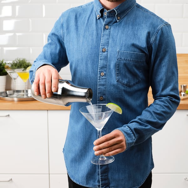 Man häller dryck från rostfritt stål shaker i cocktailglas med limeklyfta.
