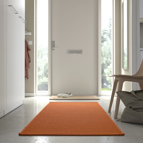 Ljusorange MORUM-matta vid dörren, passar hallar, med vita sneakers i närheten, modern minimalistisk inredning.