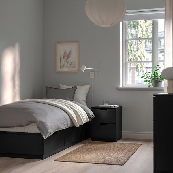 Minimalistiskt sovrum med blå väggar, svart NORDLI-säng, vita sängkläder, väggkonst, mysig belysning och ett stort fönster.