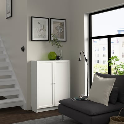 Ett litet OXBERG-skåp i vitt med grön vas står under inramad konst nära trappan, bredvid ett stort fönster med grå soffa.