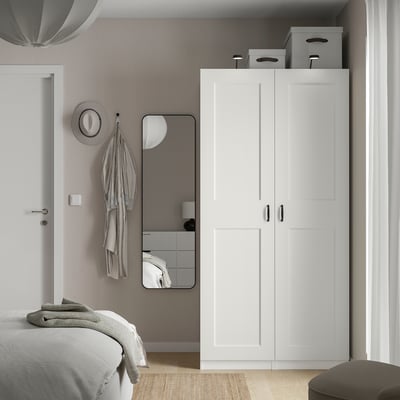 Vit PAX garderob och spegel bredvid sängen, dubbeldörrar, nätkorgar, modern design.