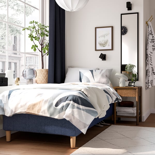 Modern sovrum med RÅHOLMAR-säng, blå lakan, nattduksbord, spegel och fönster.