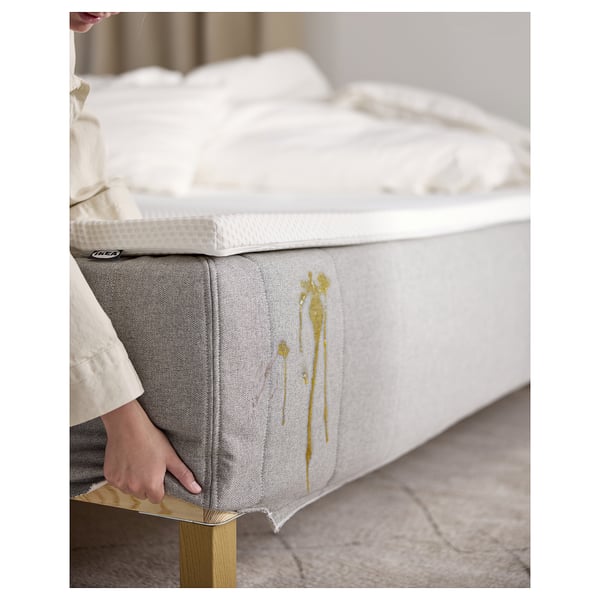 En person som justerar sängen RISEHÖJDEN , vit och grå med gulddetaljer, tillverkad av återvunnen polyester och polypropen, på en träbotten.