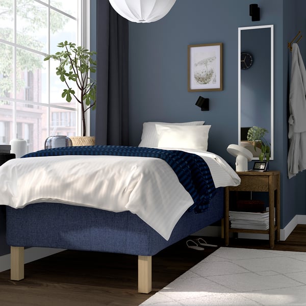 Minimalistiskt sovrum med blå RISHÖJDEN-säng, vitt täcke och blå detaljer. Mörkblå vägg med spegel, växt, nattduksbordslampa och fönster.