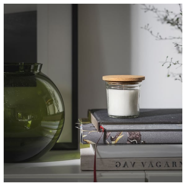Liten rund glasburk med vitt ljus och korklock står ovanpå böcker bredvid en grön vas.