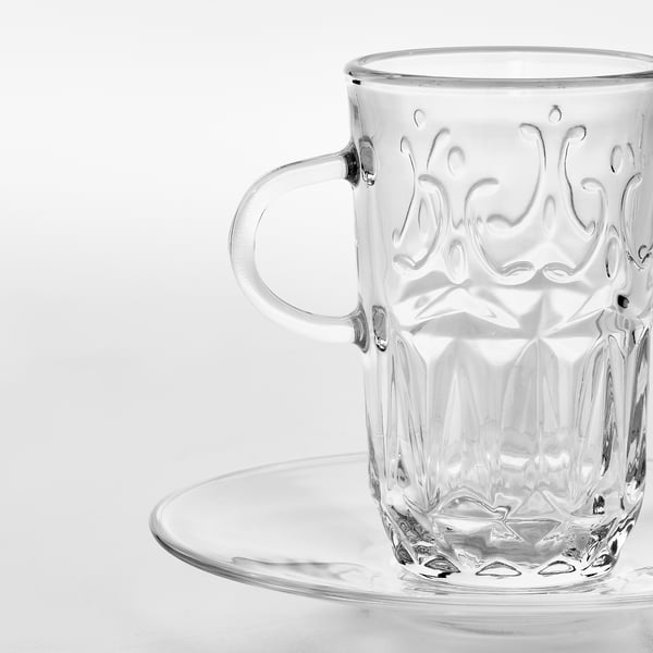 Mugg i klart glas med intrikat blommig design sitter på matchande fat. Slätt, tjockt handtag, grund kant. Elegant, tidlös.