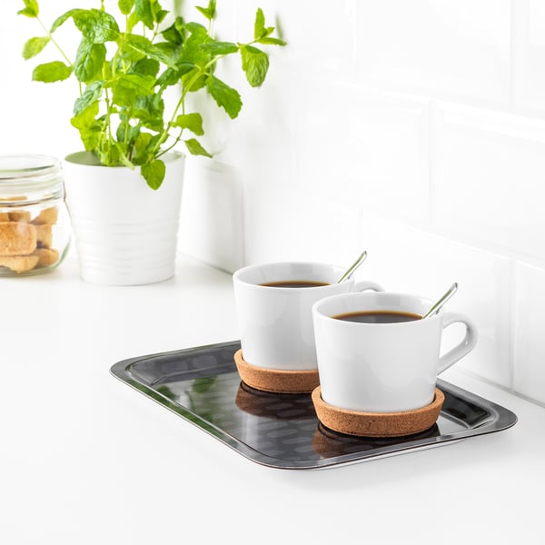 Två vita kaffekoppar med fat på en metallbricka. Gröna skedar vilar i koppar. Brickan står på en vit bänkskiva nära en växt och kakor.