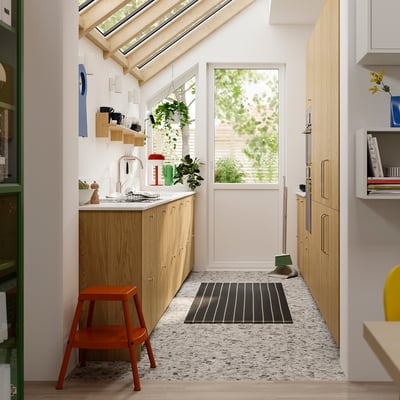 Modernt kök med ljusa träskåp, vita bänkskivor och en röd stol. Ett fönster och en dörr leder till en uteplats.