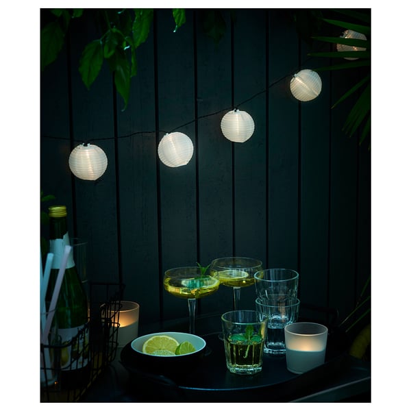 Ljusslingor lyser upp en uteplats på natten. Glas, limeskivor och stearinljus vilar på ett bord mot ett trästaket.