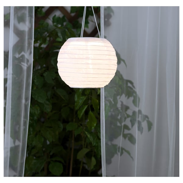 En hängande vit papperslykta lyser upp, driven av solenergi. Den är omgiven av frodig grönska och laddas under dagen och lyser upp på natten.