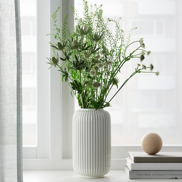 En hög vit vas med horisontella åsar håller höga gröna växter som står på en fönsterbräda. Bredvid den finns böcker och en träkula.