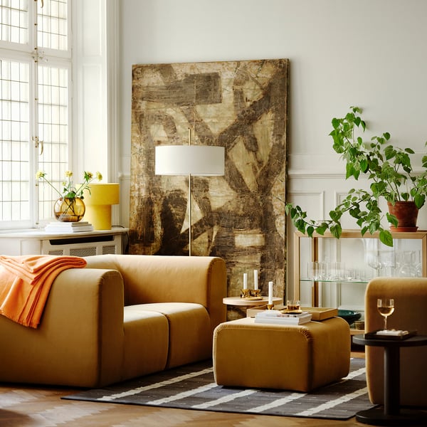Modernt vardagsrum med stor abstrakt konst, brun soffa, gul lampa och växter. Soffan är stor och fyller nästan rummet.